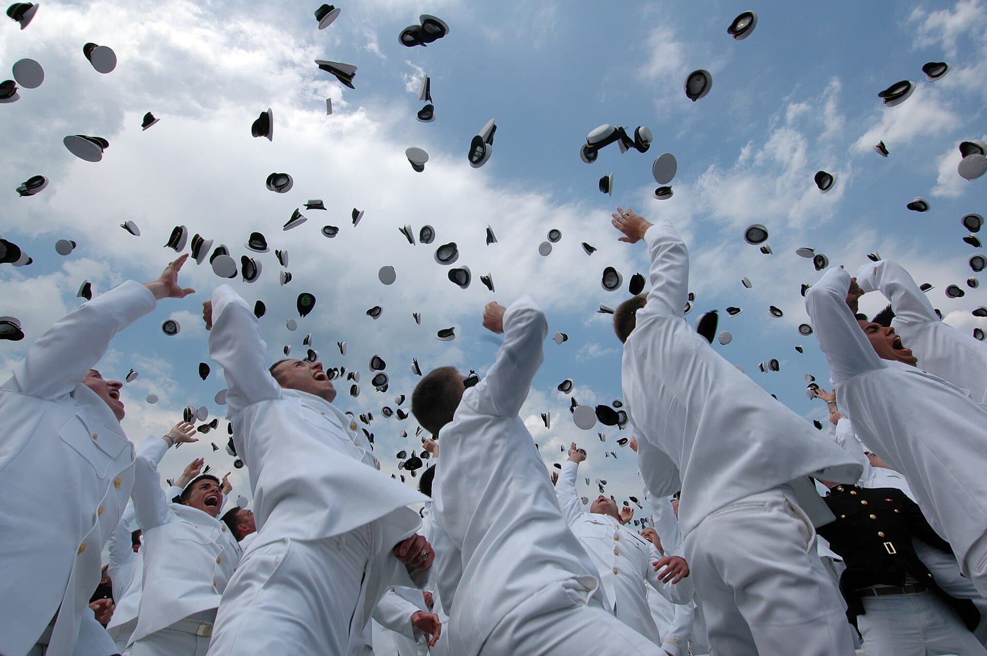 Dozens of white naval caps in the air against a blue sky, thrown by graduating midshipmen in white uniforms.