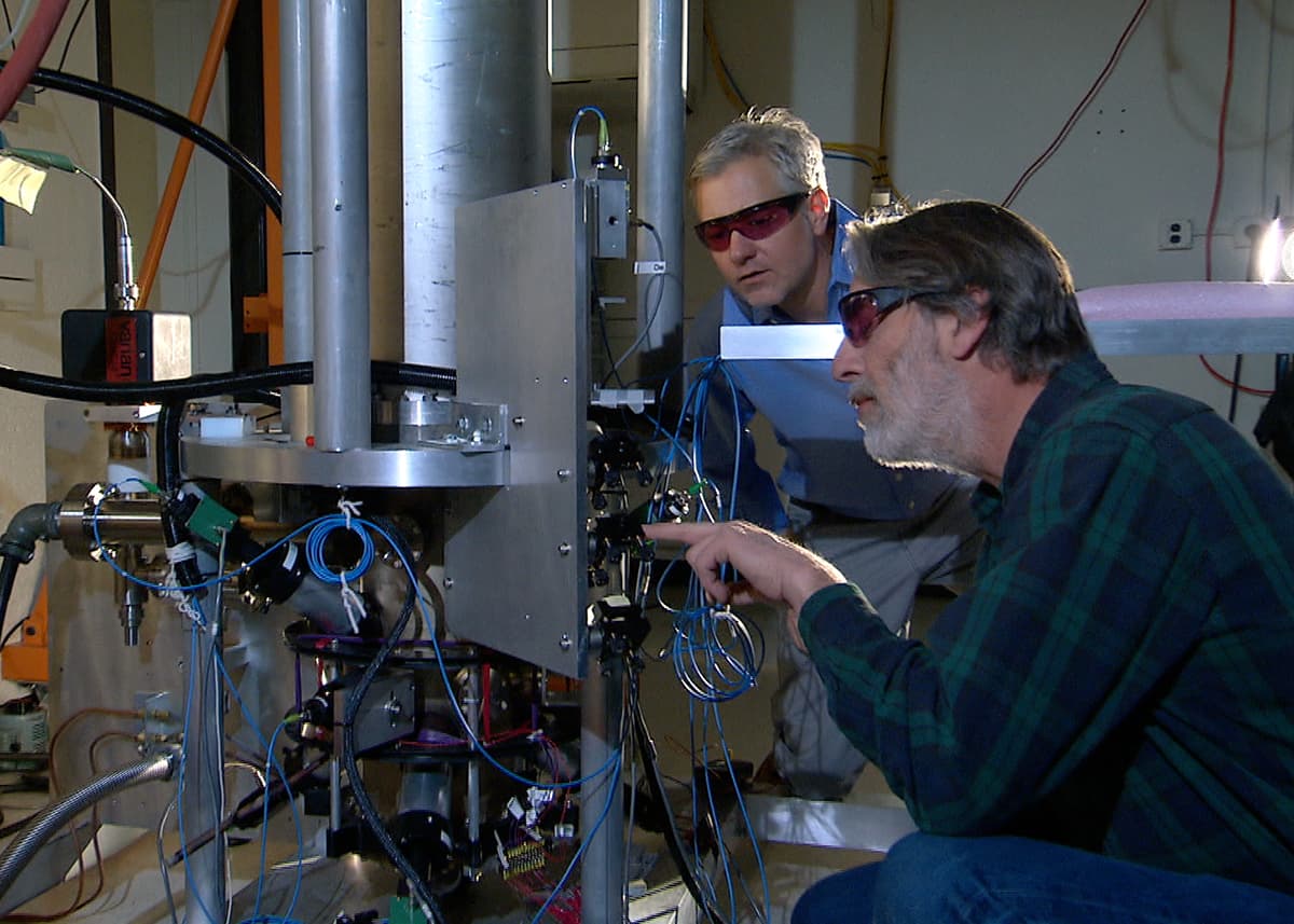 Two physicists in a laboratory beside a tall cylindrical atomic clock apparatus wrapped in cabling.